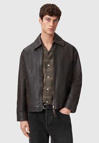 Giacca di pelle marrone con un ampio colletto, chiusura lampo frontale e finitura texture. Indossata sopra una camicia a quadri beige e nera e jeans scuri.