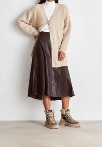 Beige fluffy cardigan over een witte coltrui, gecombineerd met een bruine leren rok en tan veterschoenen met bontafwerking en een gestructureerde zool.