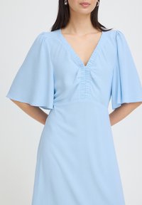 Femme portant une robe bleu clair avec des manches flottantes et un col en V froncé, debout contre un fond uni clair.