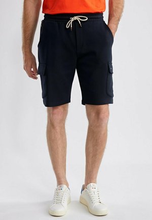 Homme portant un short cargo bleu marine avec taille à cordon, chemise orange et baskets blanches, debout devant un fond uni.