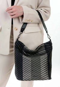 Persona in abito beige che tiene una borsa tessuta nera con texture e tracolla regolabile su uno sfondo bianco.