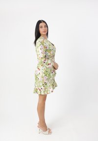 Blumenkleid in Grün mit pinken und orangefarbenen Akzenten, mit einem gerafften Saum und langen Ärmeln. Das Model trägt beiges High-Heel-Sandalen.