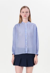 Sininen ja valkoinen raidallinen paita, jossa on pystykaulus, nappikiinnitys, pitkät hihat ja löysä istuvuus, yhdistettynä tumman väriseen laskostettuun hameeseen.