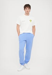 White t-shirt with a small green mushroom graphic and text; paired with light blue sweatpants; model wears white sneakers.