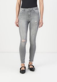 Jean skinny gris déchiré avec ourlet effiloché, présentant des accents déchirés et un design classique à cinq poches, associés à des chaussures plates noires.