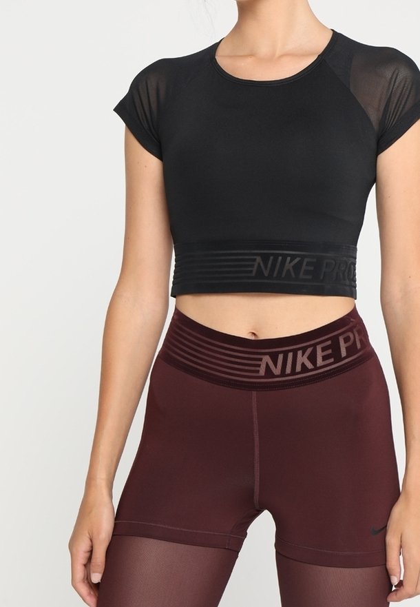 Nainen, joka on pukeutunut mustaan lyhytcroppiseen urheiluyläosaan ja viininpunaisiin korkeavyötäröisiin Nike Pro -shortseihin, joissa on läpinäkyvät leggingsit alla, seisoo yksinkertaista taustaa vasten.