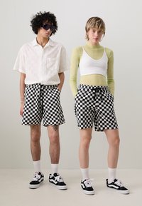 Sort-hvidternede shorts; modellen bærer en hvid skjorte med et struktureret mønster. Accessoriseret med solbriller og matchende fodtøj.