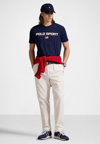 T-shirt en coton marine avec impression "POLO SPORT", pantalon crème, pull rouge noué autour de la taille, casquette marine et baskets bleues avec accents blancs.