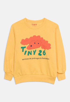 Rumena majica z dolgimi rokavi z rdečim abstraktnim obrazom, zelenim besedilom "TINY 26," in črnim francoskim besedilom spodaj spredaj.
