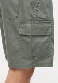 Κοντινό πλάνο πράσινων cargo σορτς με τσέπη με πτερύγιο στο πλάι και ορατή ραφή, φορεμένα από άτομο με εκτεθειμένο πόδι.