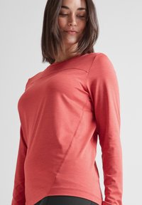 Woman looking down wearing a coral long-sleeve fitted athletic top against a plain light background.