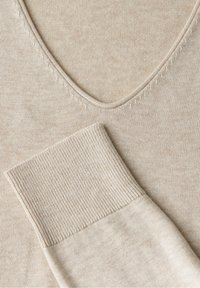 Pull beige avec un décolleté en V et des poignets côtelés, fabriqué en tissu doux avec une texture lisse et des détails de couture subtils.