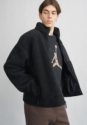 Overmaat zwart fleece jacket met een hoge kraag en ritsluiting. Voorzien van zijzakken en een gestructureerde stof. Gedragen over een zwart graphic t-shirt.