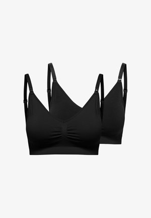 Fekete ujjatlan felsőt és magas derekú leggingset viselő terhes nő, mezítláb áll egy sima világos háttér előtt, lefelé néz.