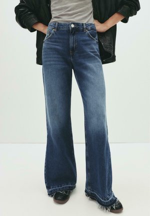 Wide-leg blue jeans with frayed hems worn with black shoes, paired with a light top and black leather jacket, hands resting on hips.