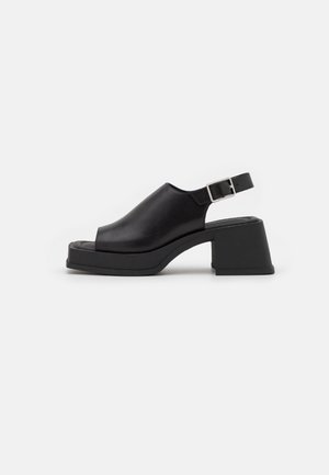Sandale slingback en cuir noir avec bout ouvert, talon moyen épais et bride à boucle argentée sur fond blanc.