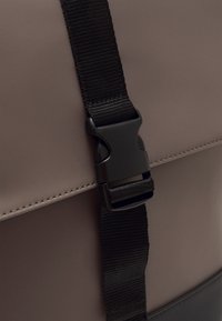 Sac à dos marron imperméable avec surface texturée, équipé d'une sangle noire réglable et d'une fermeture à boucle en plastique. Accent gris en bas.