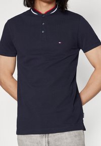 Man som bär en marinblå kortärmad pikétröja med en vit, röd och marinrandig krage samt en liten flagglogotyp på bröstet.