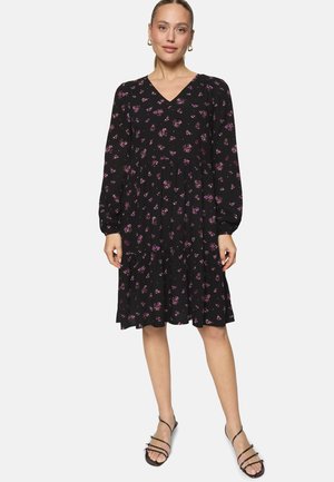 Femme portant une robe noire à manches longues, longueur genou, avec un petit imprimé floral violet et des sandales noires à brides, debout devant un fond blanc.