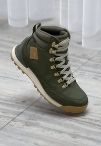 Botte de randonnée verte avec un dessus en tissu texturé, semelle en caoutchouc, œillets métalliques et lacets gris. Présente une pièce de logo sur le côté.