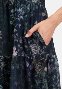 Robe fleurie en tissu bleu foncé avec une poche, des motifs botaniques complexes en plusieurs couleurs et des détails de volants superposés.
