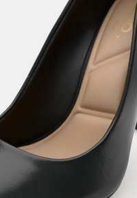 Scarpa con tacco alto in vernice nera, con una finitura liscia e una morbida fodera interna beige, caratterizzata da una silhouette curva e una superficie lucida.