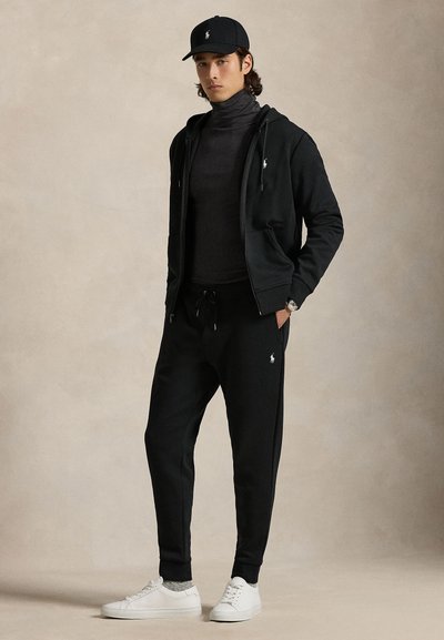 Junger Mann mit schwarzer Mütze, schwarzem Kapuzenpullover mit Reißverschluss, dunklem Rollkragenpullover, schwarzen Jogginghosen, grauen Socken und weißen Turnschuhen steht vor einem einfarbigen Hintergrund.