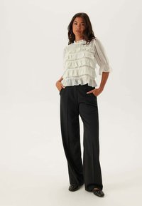 Camicia bianca con volant a tre strati, maniche lunghe a palloncino e collo alto, abbinata a pantaloni neri larghi e ballerine nere.