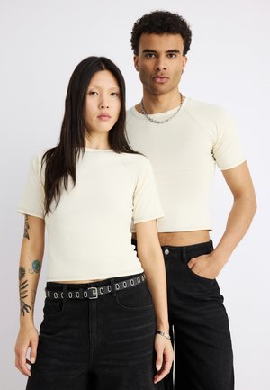 Dwie osoby w kremowych krótkich koszulkach i czarnych spodniach stoją na jednolitym białym tle, zwrócone przodem z neutralnymi wyrazami twarzy.