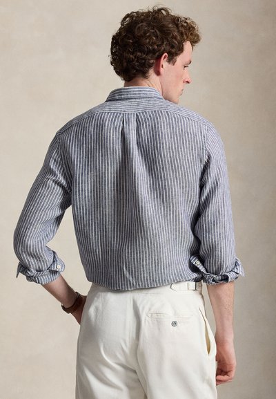 Άνδρας με σγουρά μαλλιά που φοράει μπλε και λευκό πουκάμισο με κάθετες ρίγες και μανίκια που είναι γυρισμένα προς τα πάνω, καθώς και λευκό παντελόνι, με την πλάτη του προς την κάμερα.