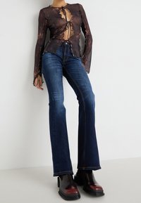 Top marrón transparente con lazos, que presenta un escote en V profundo y mangas largas; combinado con jeans de mezclilla azul de pata de elefante y botas negras hasta el tobillo con acentos rojos.