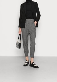Zwarte blazer over een zwarte coltrui, grijs geruite broek en zwart-witte schoenen. Kleine zwarte handtas met kettingband.