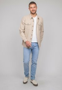 Chemise beige à boutons avec deux poches poitrine, portée sur un t-shirt blanc, assortie à un jean bleu clair et des chaussures slip-on beiges.