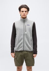 Νέος άνδρας που φοράει γκρι γιλέκο φλις με φερμουάρ πάνω από μαύρο μακρυμάνικο πουκάμισο και λαδί cargo σορτς, στέκεται μπροστά σε απλό φωτεινό φόντο.