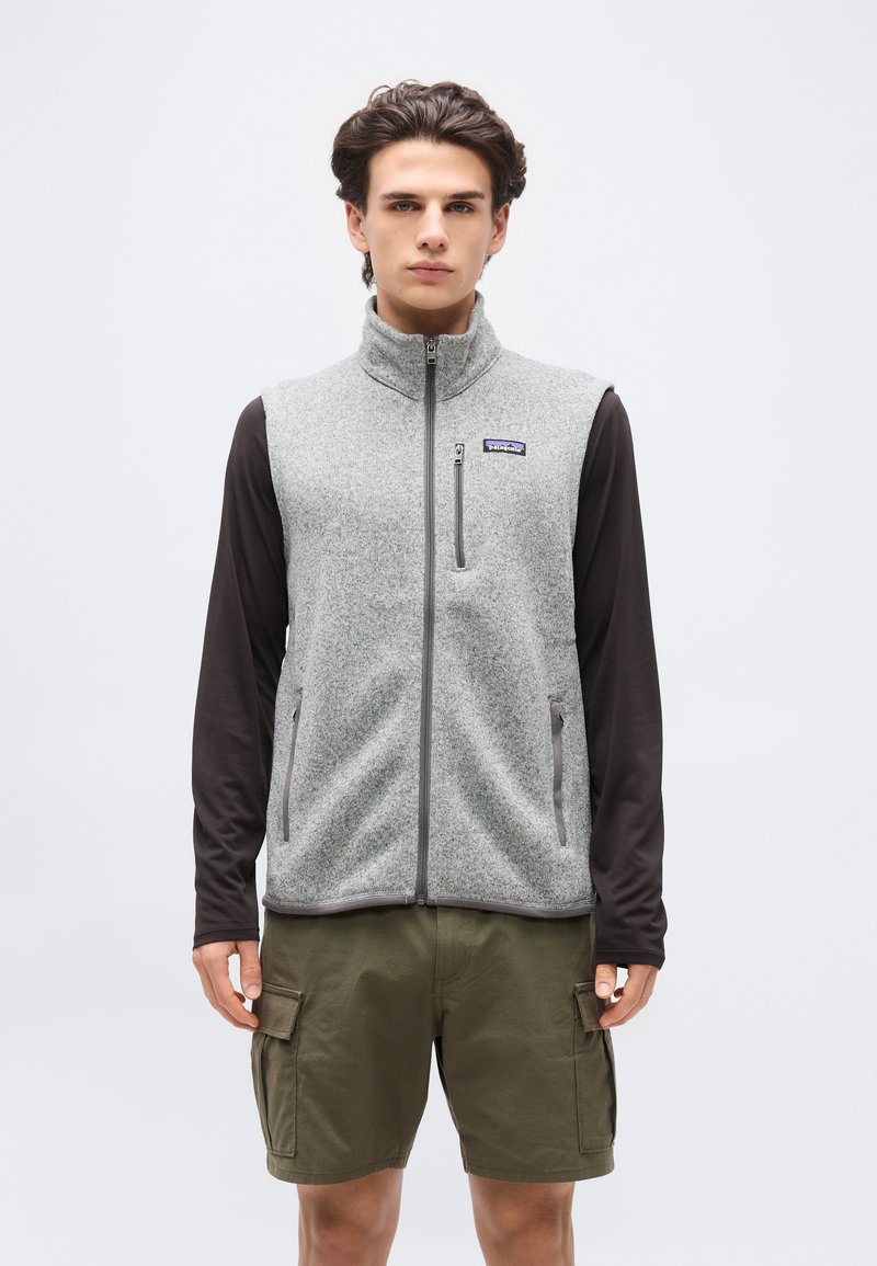 Νέος άνδρας που φοράει γκρι γιλέκο φλις με φερμουάρ πάνω από μαύρο μακρυμάνικο πουκάμισο και λαδί cargo σορτς, στέκεται μπροστά σε απλό φωτεινό φόντο.