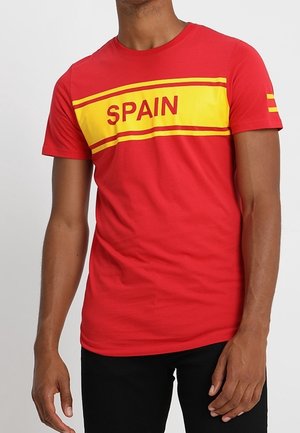 Mężczyzna w czerwonej koszulce z żółtym paskiem i napisem "SPAIN" na klatce piersiowej, w czarnych spodniach.