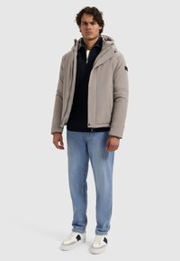 Lichtgrijze pufferjack met capuchon, ritssluiting en zwarte zakdetails, gedragen boven een marineblauw trui en lichtblauwe jeans, gecombineerd met witte sneakers.