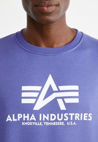 Vijolična športna bunda z rebrastim okroglim ovratnikom, ki vsebuje bel grafični dizajn, vključno z zvezdo in besedilom "ALPHA INDUSTRIES, KNOXVILLE, TENNESSEE, ZDA."