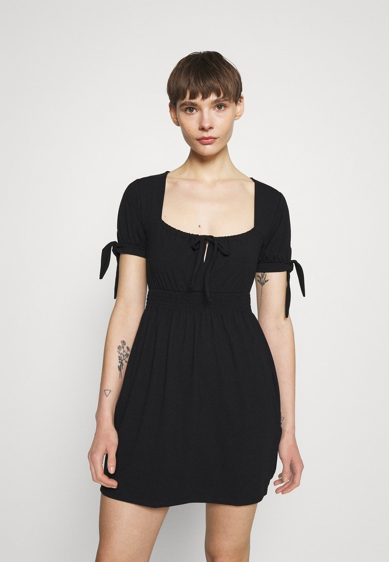 Vestido preto curto com decote quadrado, mangas bufantes presas com laços, cintura elástica e tecido liso. Apresenta detalhes franzidos sutis.