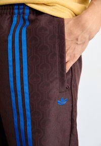 Pantaloni di felpa marroni con motivo, caratterizzati da strisce blu ai lati e un logo ricamato blu. Il tessuto appare liscio con un leggero lucido.