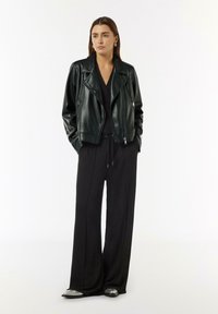 Czarna skórzana kurtka motocyklowa o dopasowanym kroju, z zamkiem błyskawicznym z przodu i klapami, noszona na czarnym swetrze i szerokich spodniach.