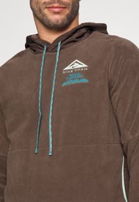 Felpa in pile marrone con cordino turchese ed emblema ricamato. Il design include una grafica di montagna e del testo.