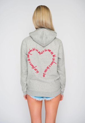 Hoodie gris avec une capuche à cordon, présentant un texte rouge en forme de cœur au dos. Le tissu est doux avec une coupe décontractée. Porté avec un short en denim.
