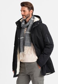 Parka nera con cappuccio foderato in pelliccia, abbinata a una sciarpa grigia a spina di pesce. Sotto, una camicia beige e pantaloni cargo completano il look.