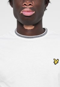 T-shirt en coton blanc avec un col à rayures noires et blanches, arborant un petit logo d'aigle jaune et noir sur la poitrine. Texture lisse.