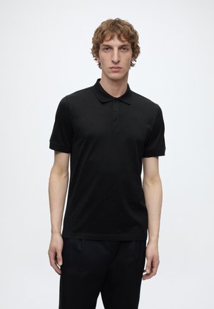 Young man with curly hair wearing a black short-sleeve polo shirt and black pants against a plain light background.