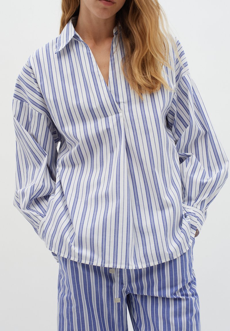 Camicia a righe bianca e blu, con maniche lunghe, colletto e vestibilità morbida. Abbinata a pantaloni a righe coordinati.