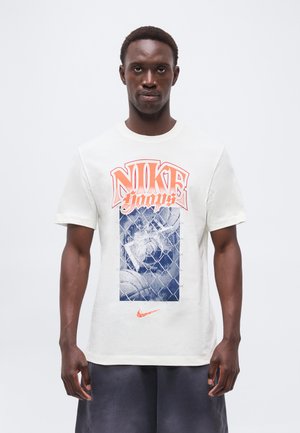 Férfi fehér Nike pólóban, narancssárga "Nike Hoops" felirattal és kék kosárlabda grafikával egy láncszemes kerítés mögött.