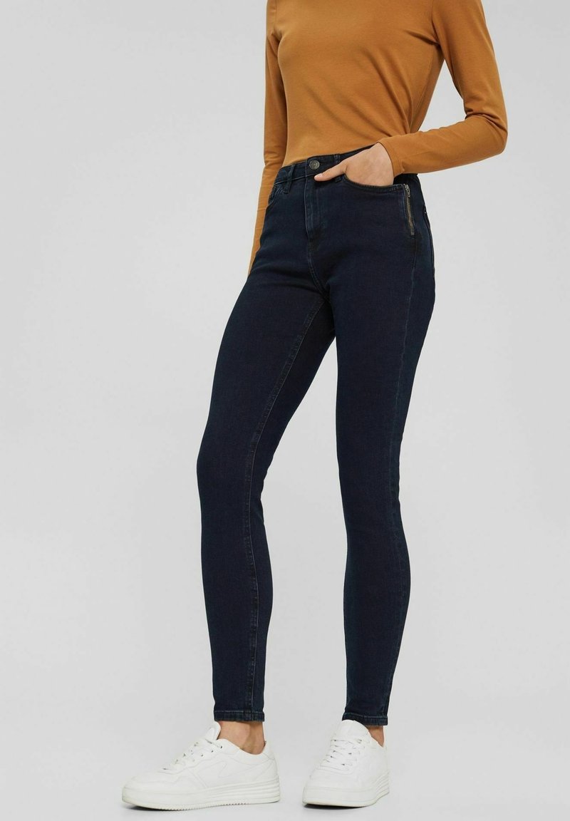 Person med mörkblå smala jeans, senapsgul långärmad tröja, vita sneakers, med en hand i framtill på fickan mot en neutral bakgrund.