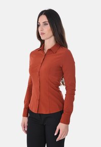 Blusa rustica a maniche lunghe, con frontale a bottoni, colletto e vestibilità sartoriale, abbinata a pantaloni neri. Texture liscia e design pulito.